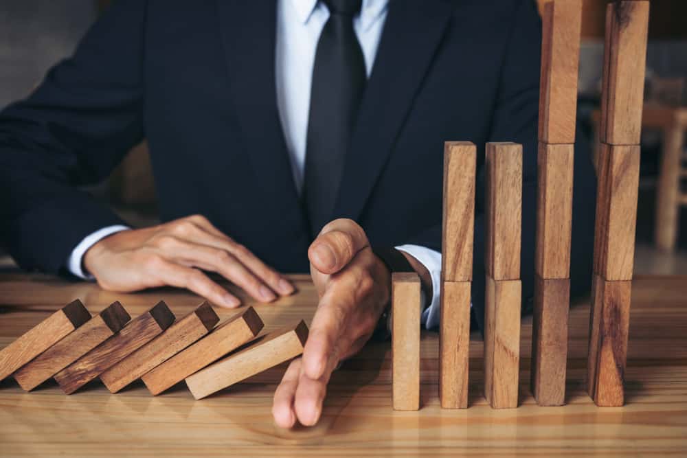 Close up of businessman hand Stopping Falling wooden Dominoes effect from continuous toppled or risk, strategy and successful intervention concept for business. Source: shutterstock.com